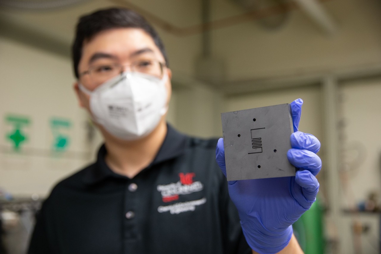 Wearing a face mask and rubber gloves, Jingjie Wu holds up catalyst reactor in a lab.