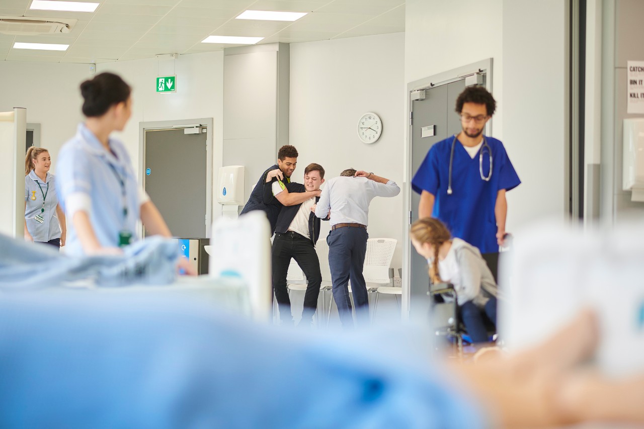 Image shows a security guard helping to subdue a visitor in an emergency room setting. Healthcare workers stand nearby.